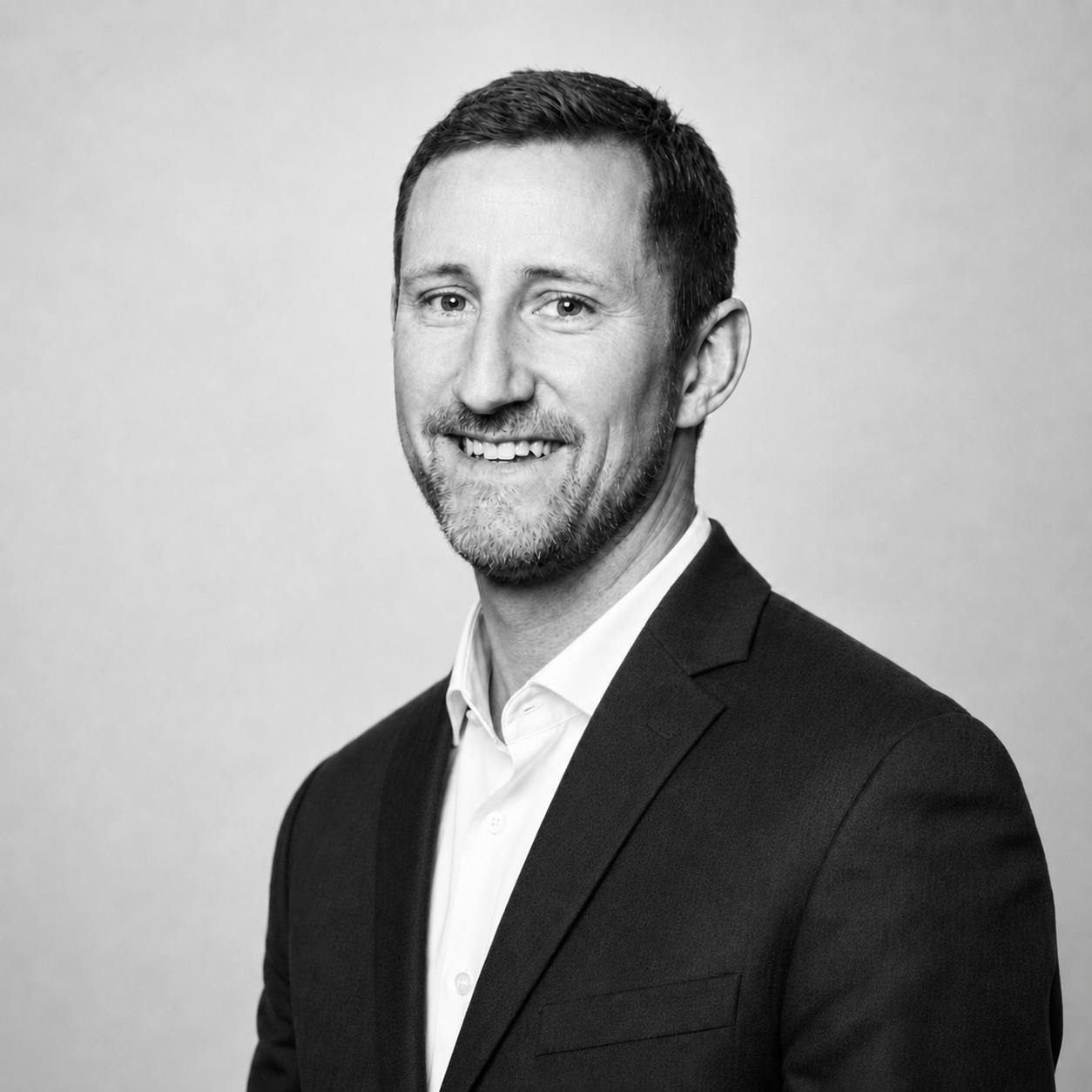 Black and white professional headshot of a smiling man with short hair and a beard, wearing a suit jacket and collared shirt, against a plain light background.