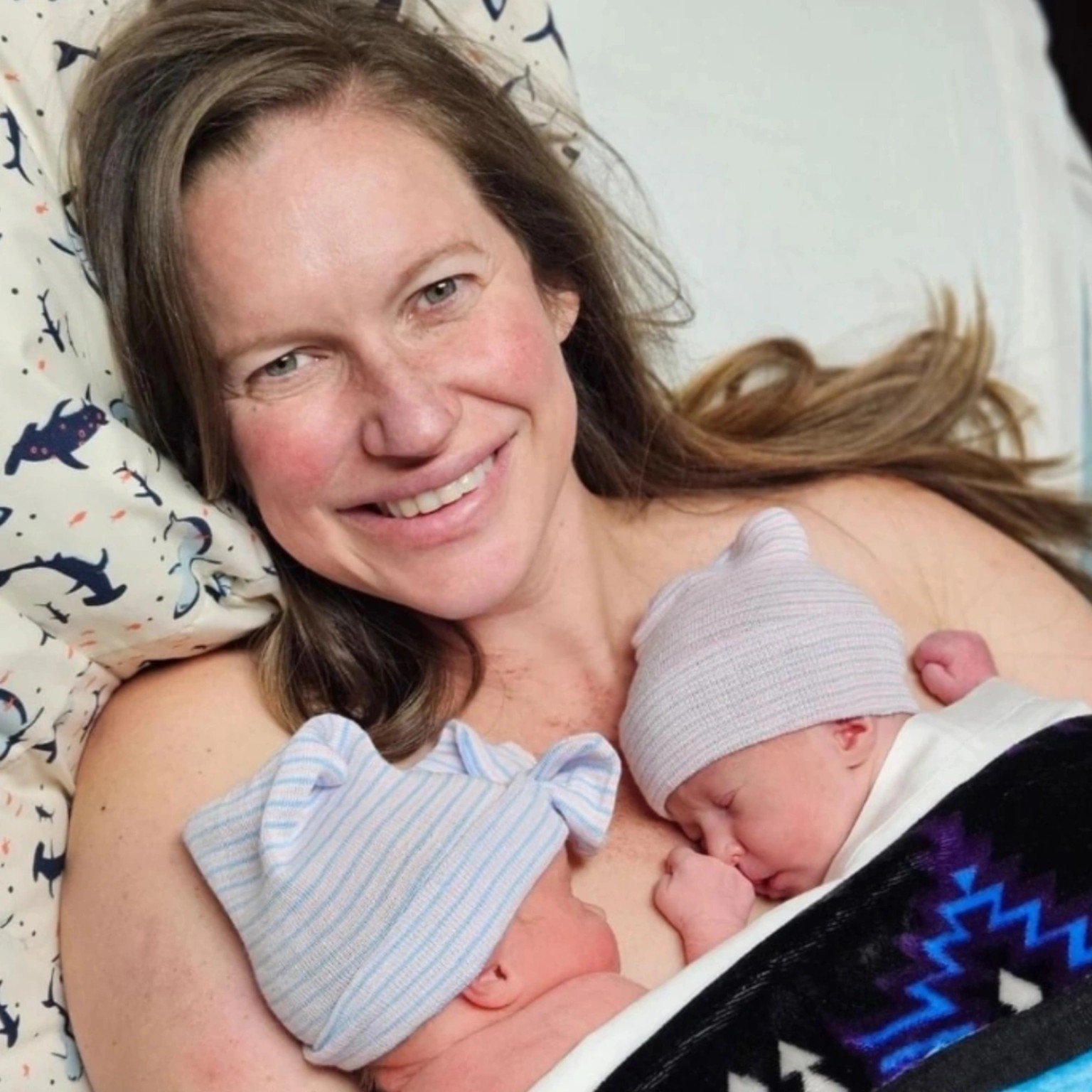 A woman with long brown hair lying on a bed, smiling, while breastfeeding two babies, one dressed in a striped shirt and the other in a white shirt with a beanie.