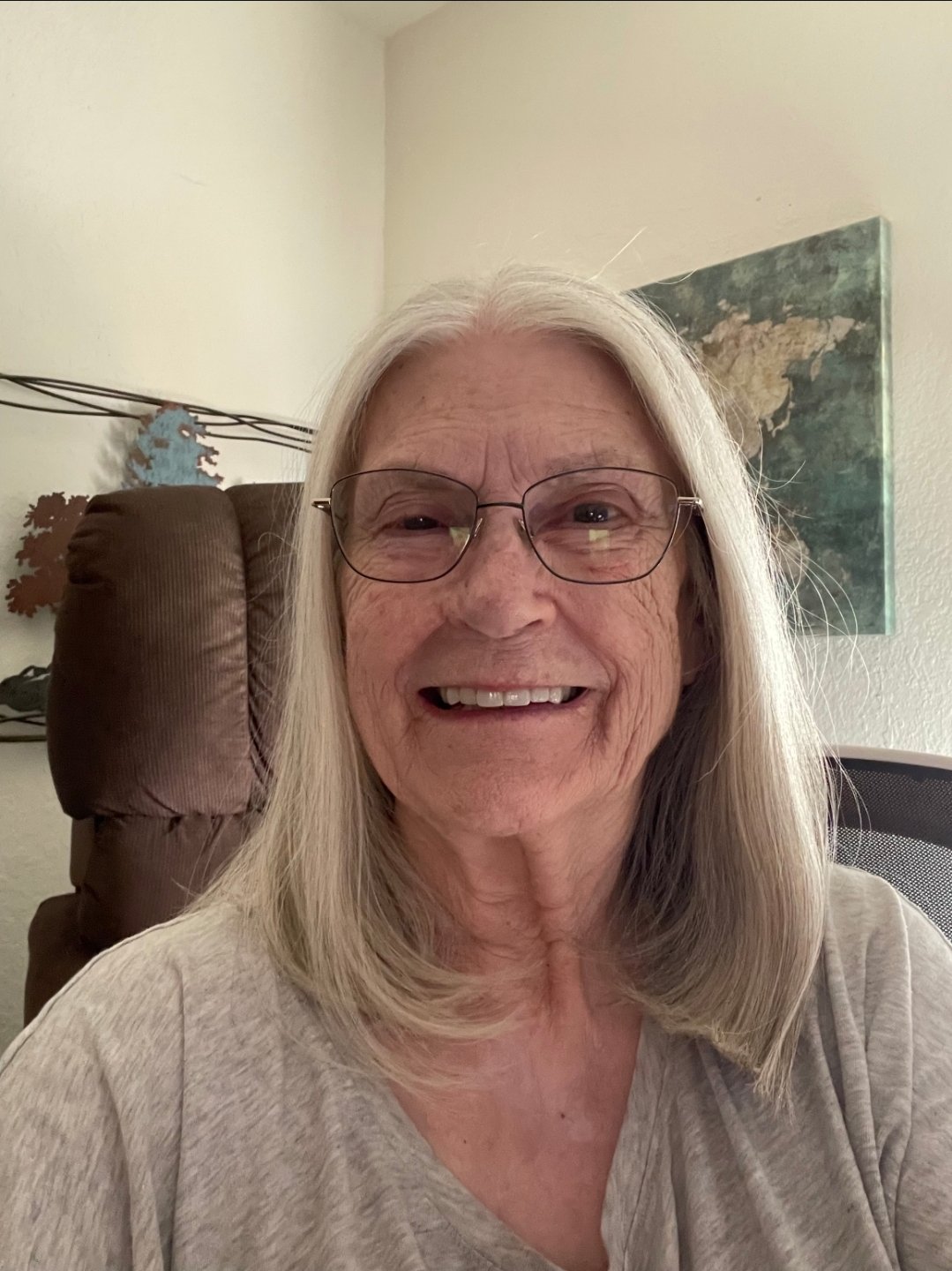 A smiling elderly woman with long gray hair, glasses, and a beige shirt, sitting in a room with a world map on the wall behind her.