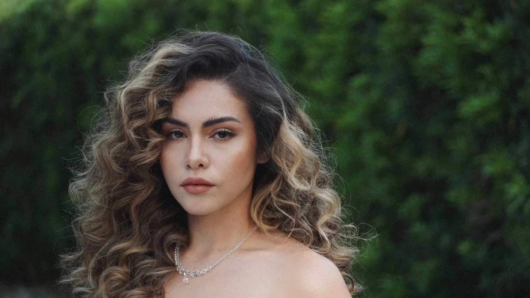 A portrait of a woman with curly, voluminous hair, wearing a diamond necklace, against a background of greenery.