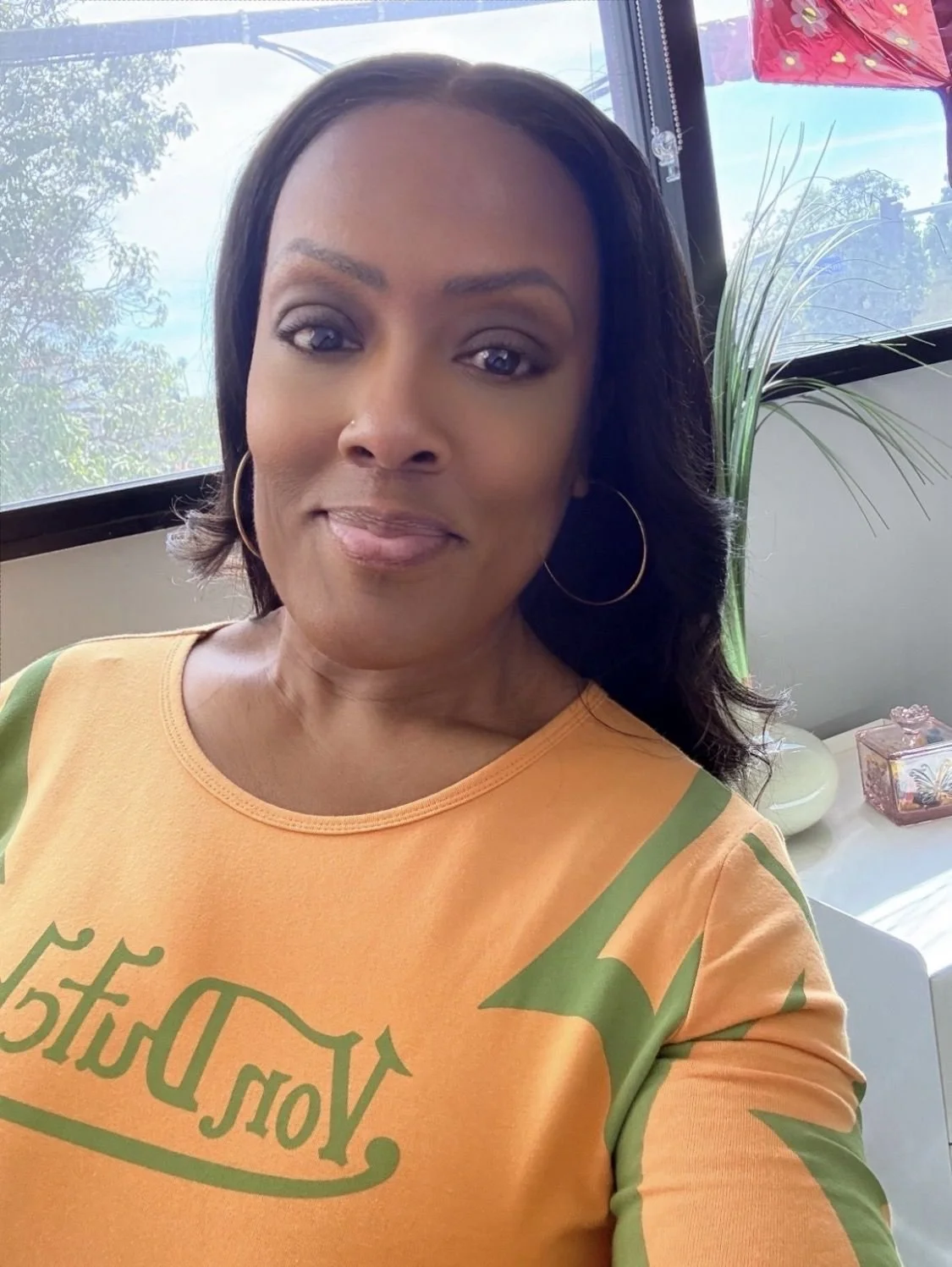 A woman with shoulder-length dark hair and hoop earrings smiling at the camera, wearing an orange and green shirt, indoors near a window with trees outside and a plant behind her.