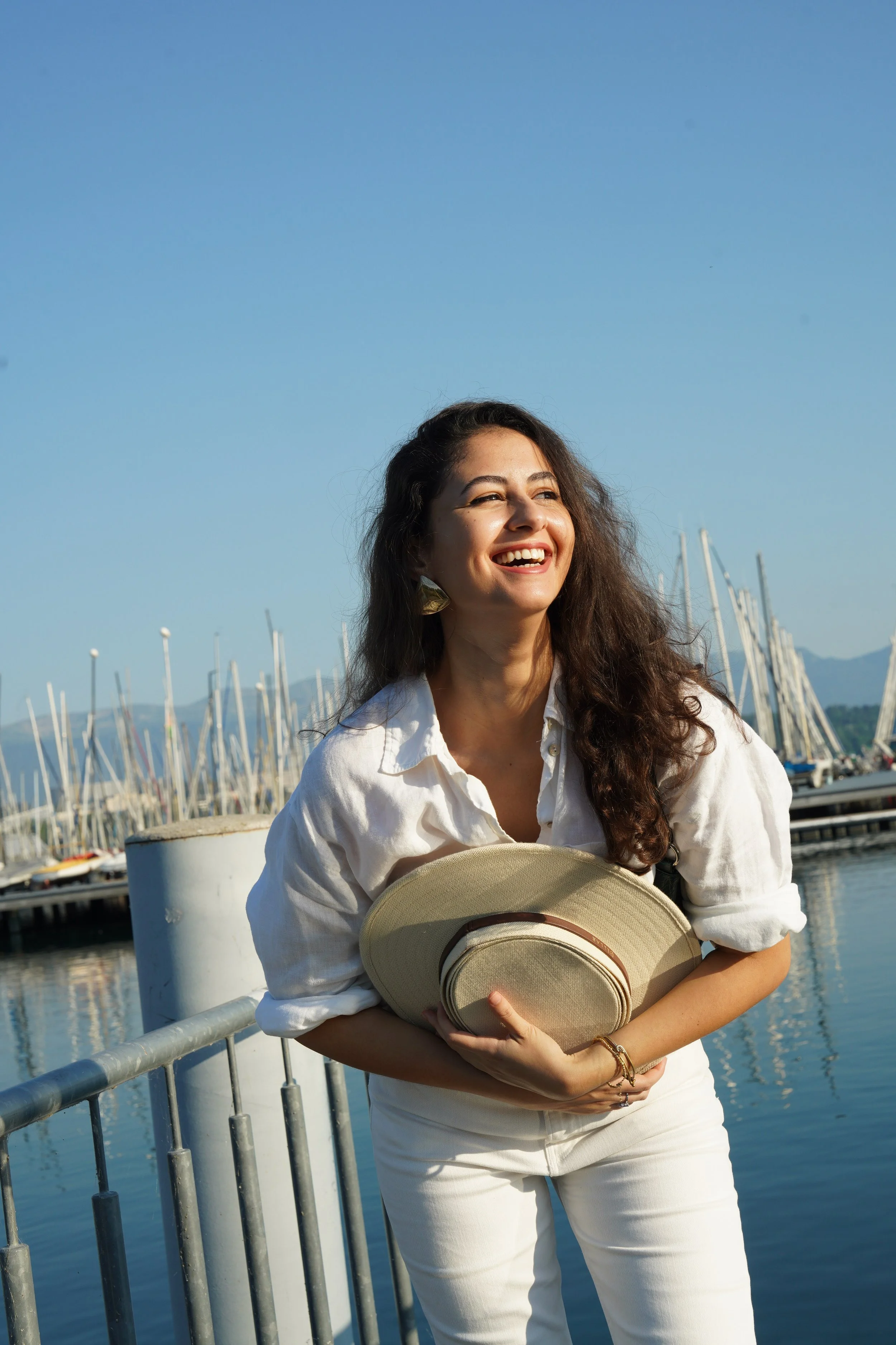 A woman with long, curly brown hair smiling and holding a beige hat in front of a marina with sailboats docked and a blue sky.