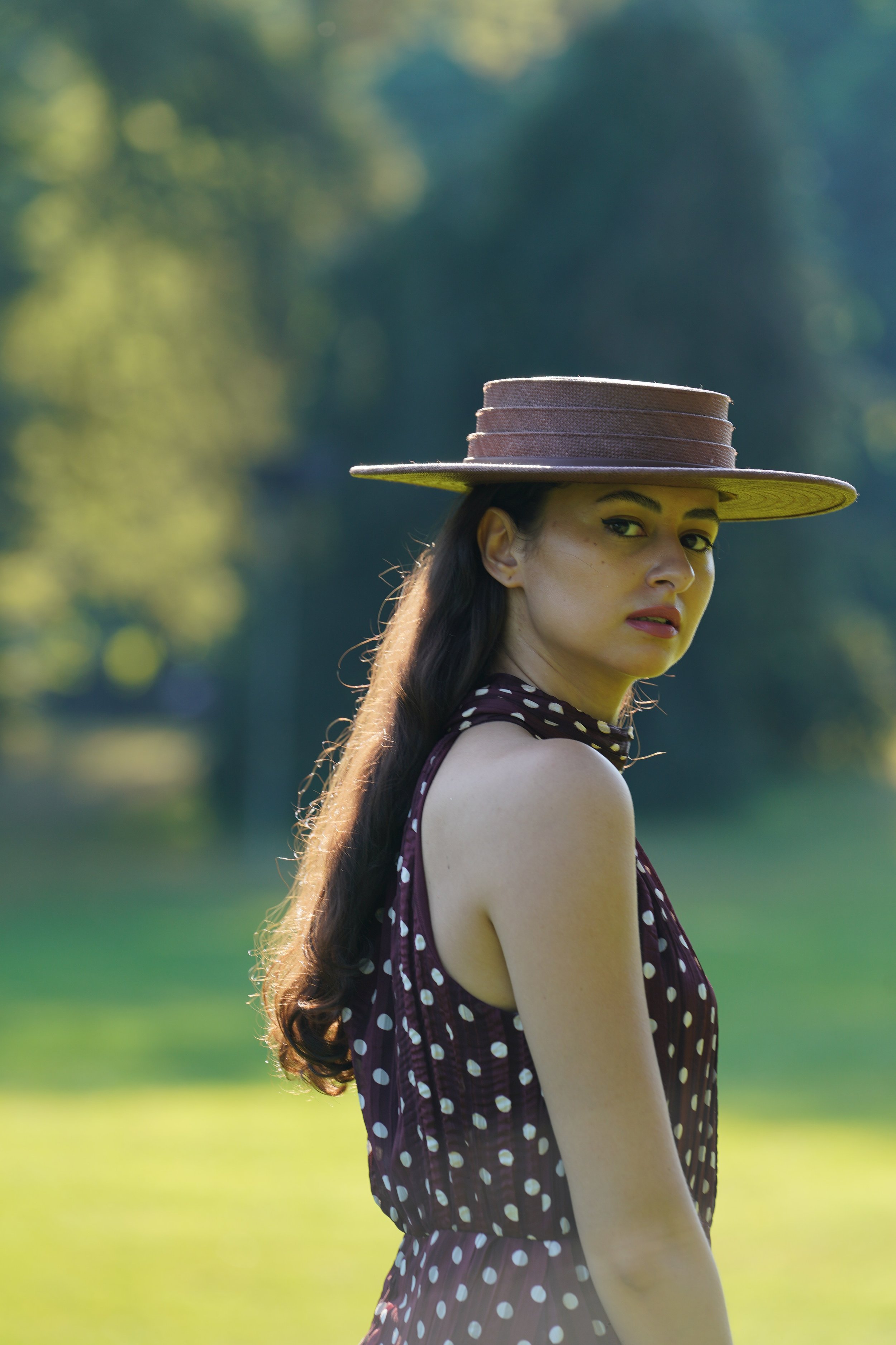A woman with long brown hair wearing a wide-brimmed straw hat and a sleeveless maroon polka-dot dress standing outdoors in a park with trees and grass in the background, looking over her shoulder.