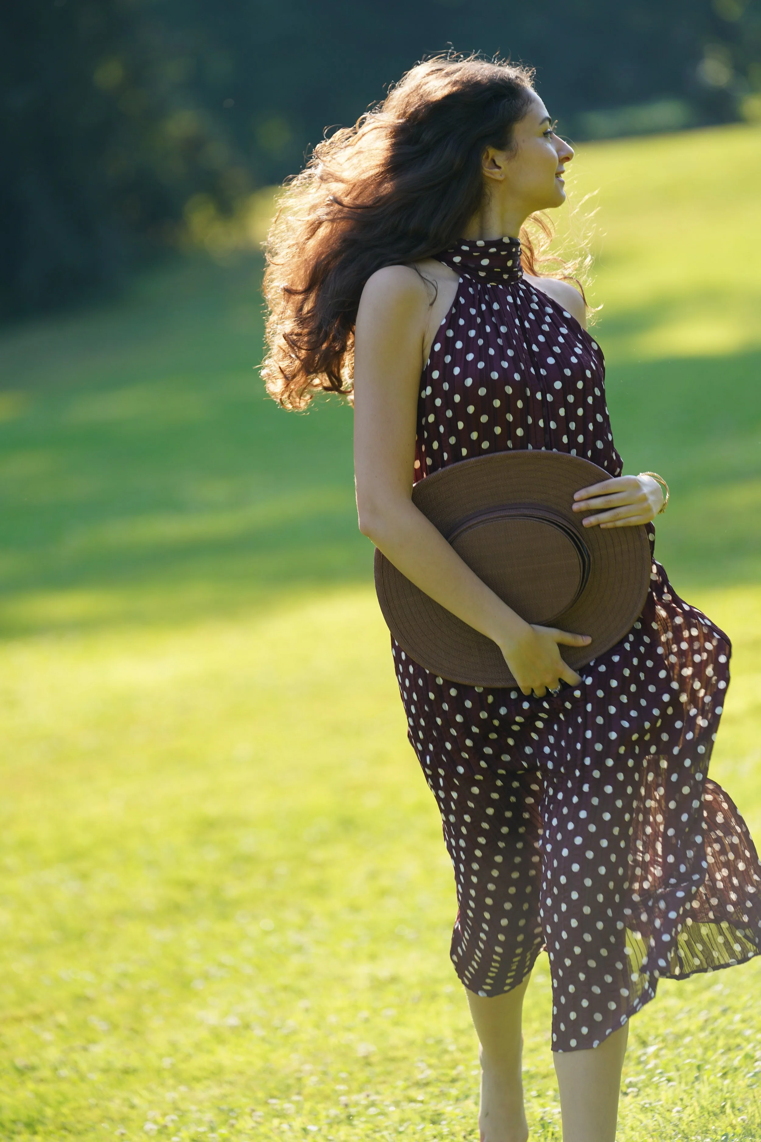 A woman with long curly hair stands outdoors in a grassy area, holding a wide-brimmed hat, wearing a sleeveless polka dot dress, smiling and looking to the side with sunlight illuminating her face and hair.