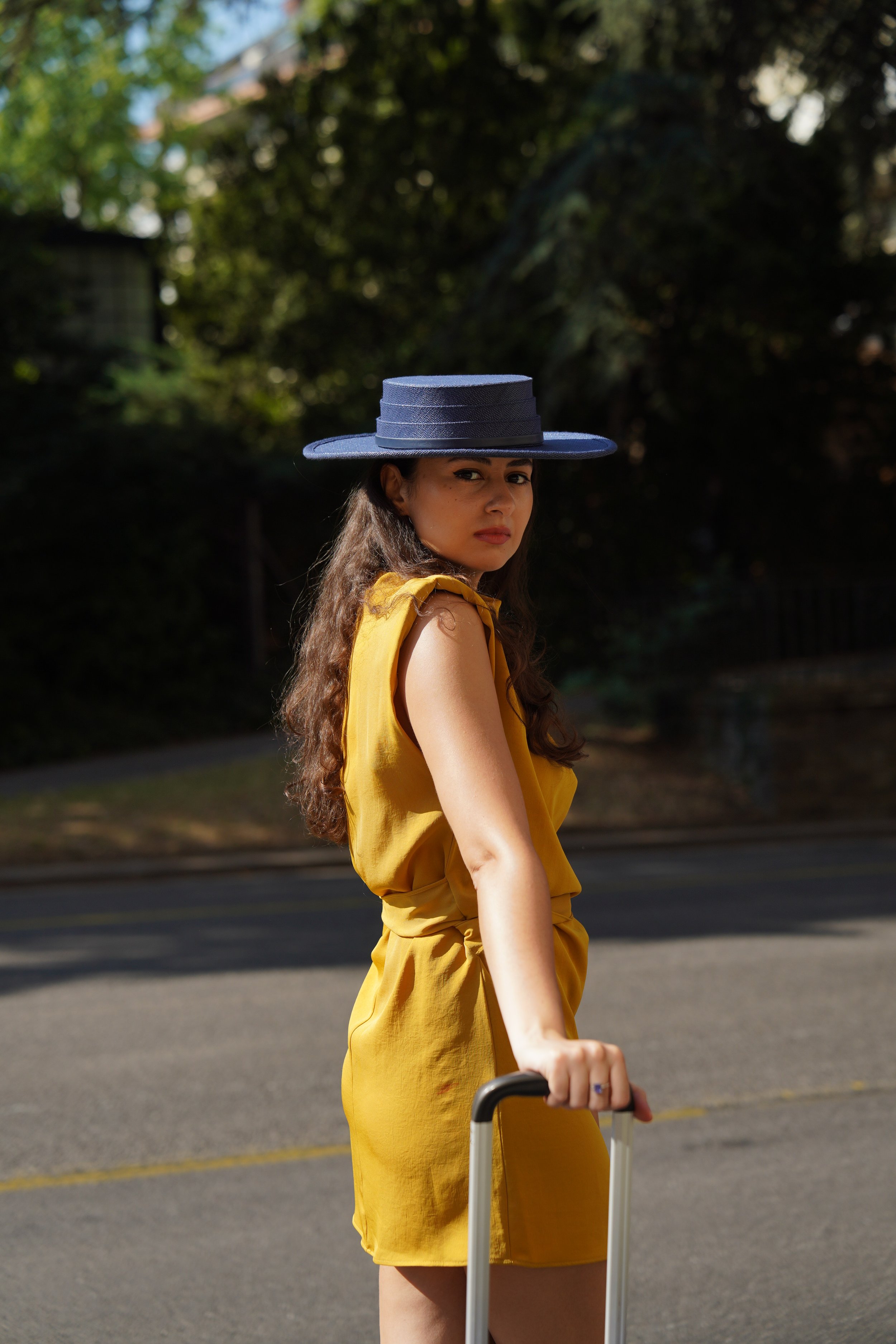 Young woman with long brown hair wearing a large navy blue hat and a yellow outfit, standing outdoors near a street.