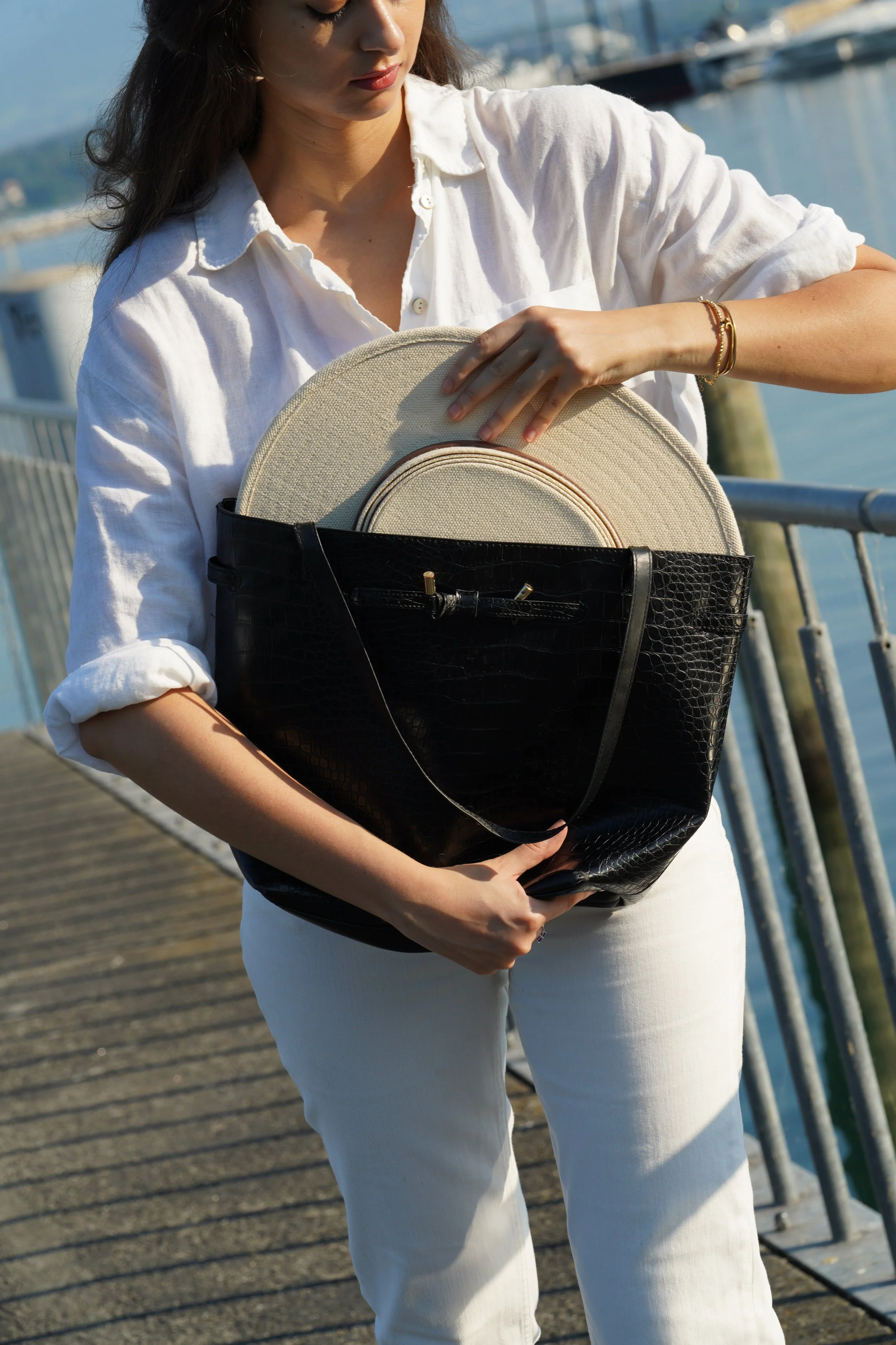 A woman in a white shirt holding a beige hat and a black handbag on a wooden pier with water and boats in the background.