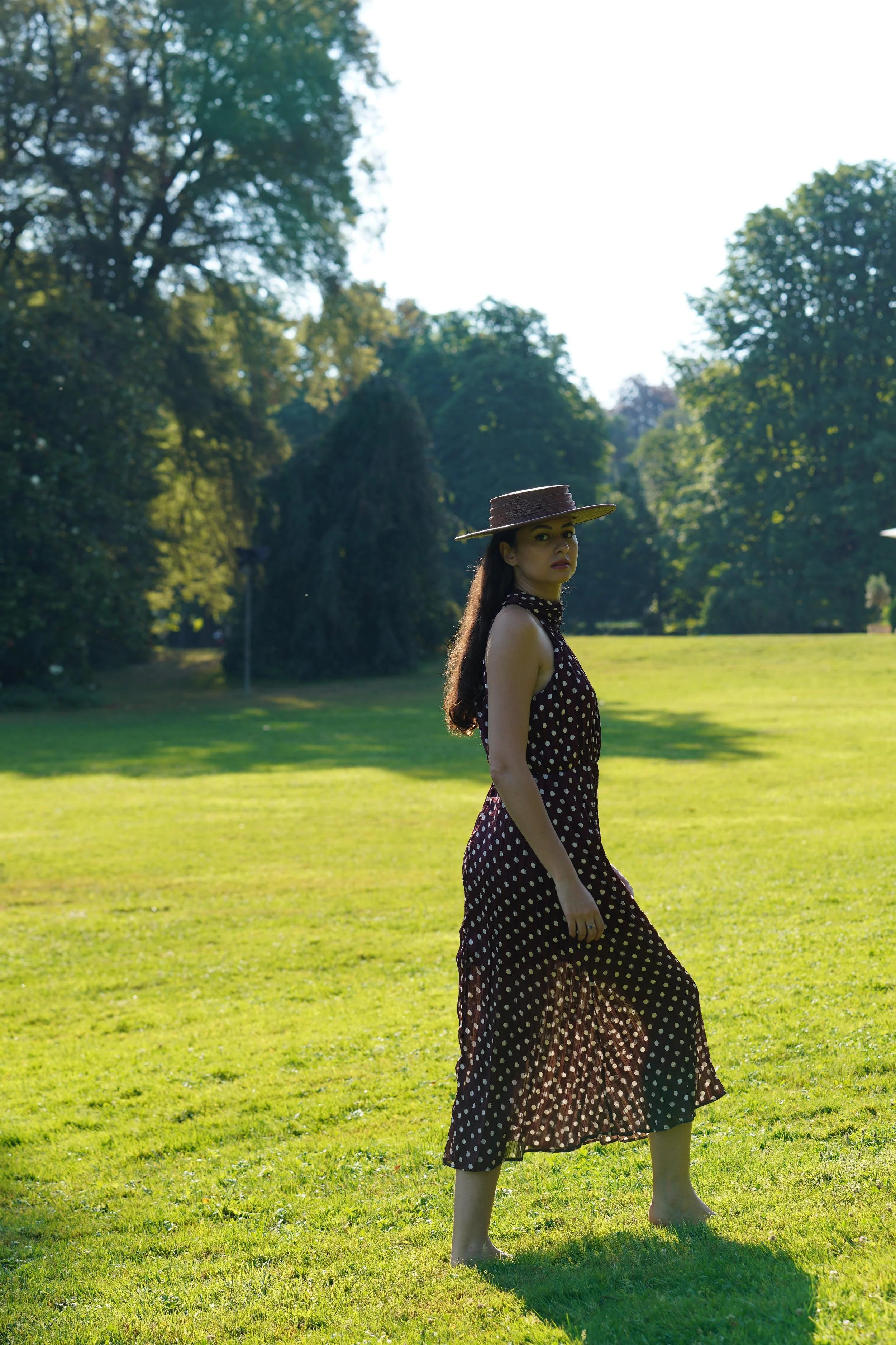 A woman with long dark hair wearing a wide-brimmed brown hat and a vintage polka dot dress stands barefoot on a grassy field in a park with tall trees and clear sky in the background.