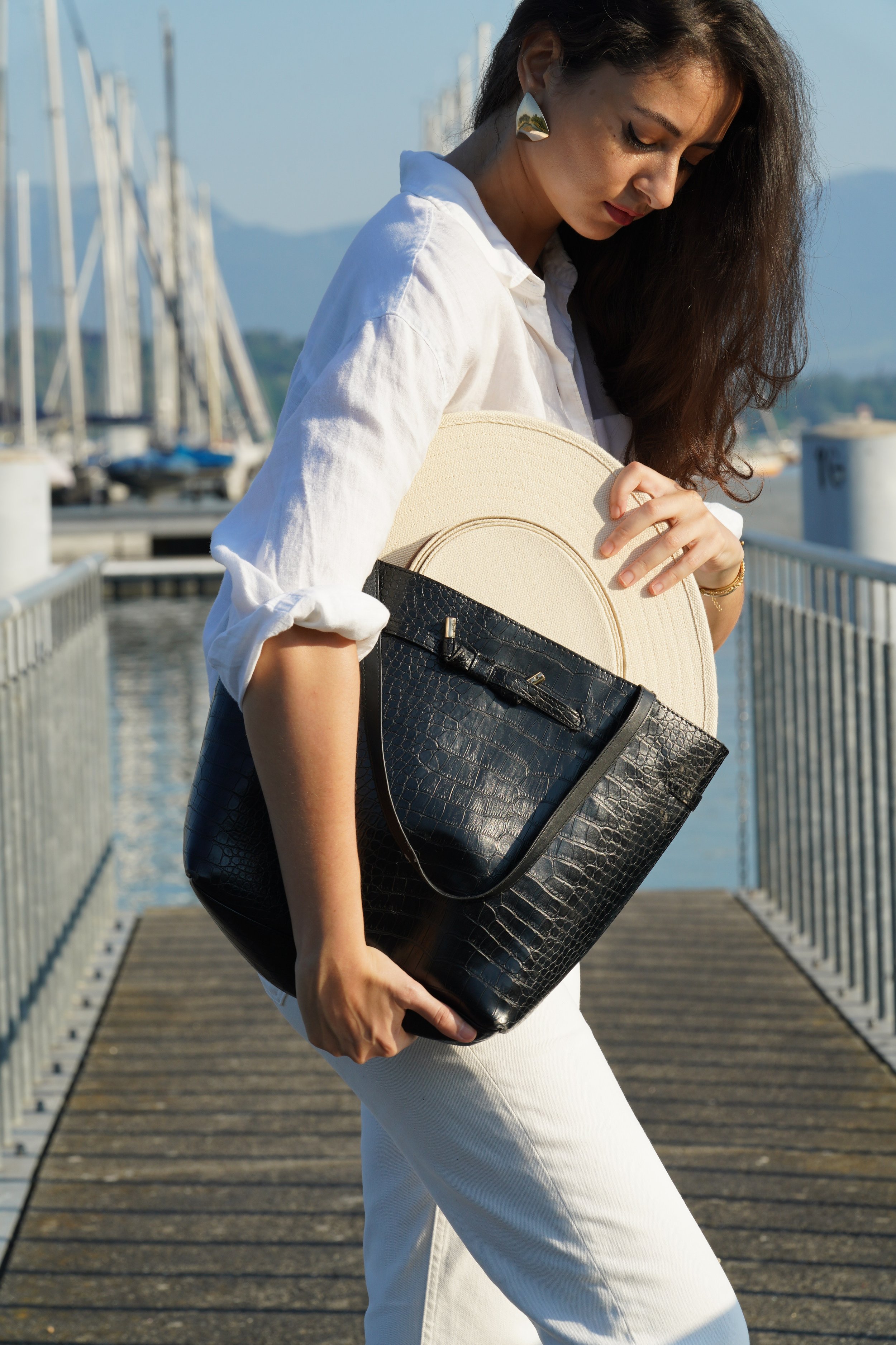 A woman with long, wavy brown hair, wearing a white shirt and white pants, holding a large black and beige purse, standing on a wooden dock near the water with sailboats in the background on a sunny day.