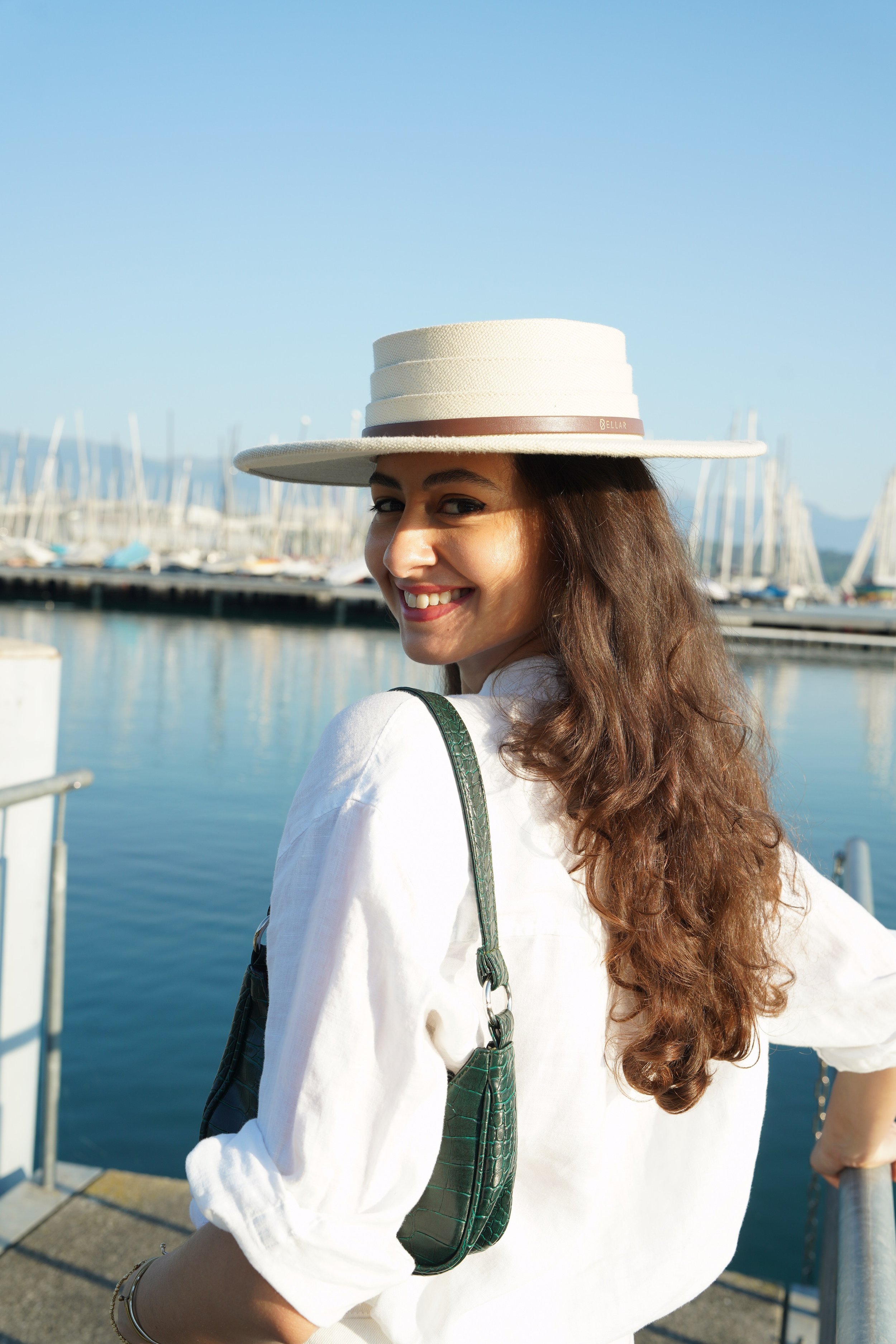 A woman with long curly hair and a wide-brimmed hat smiling on a boat dock with sailboats in the background.