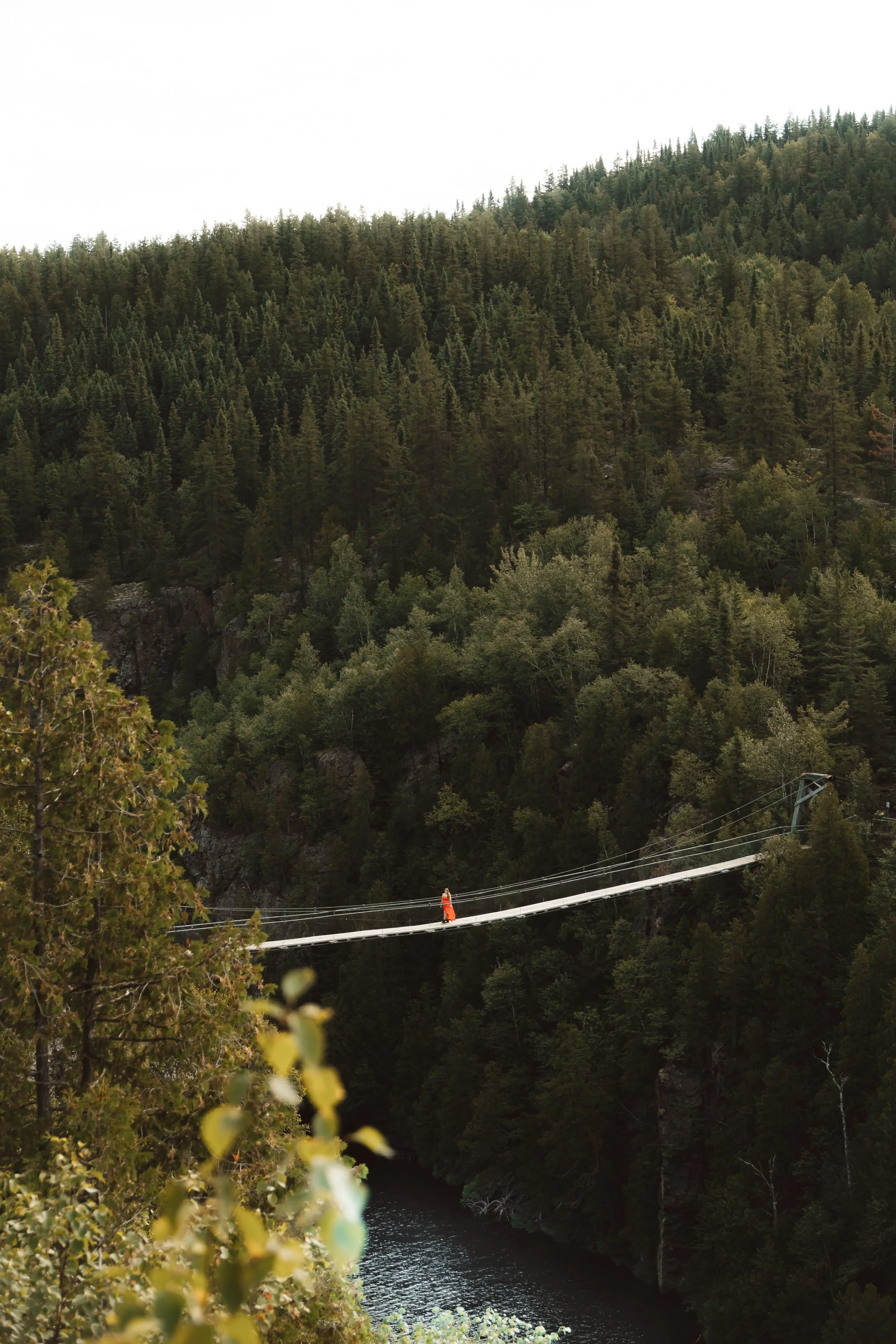 A person in a red dress crossing a suspension bridge over a river in a dense forested mountainous area.
