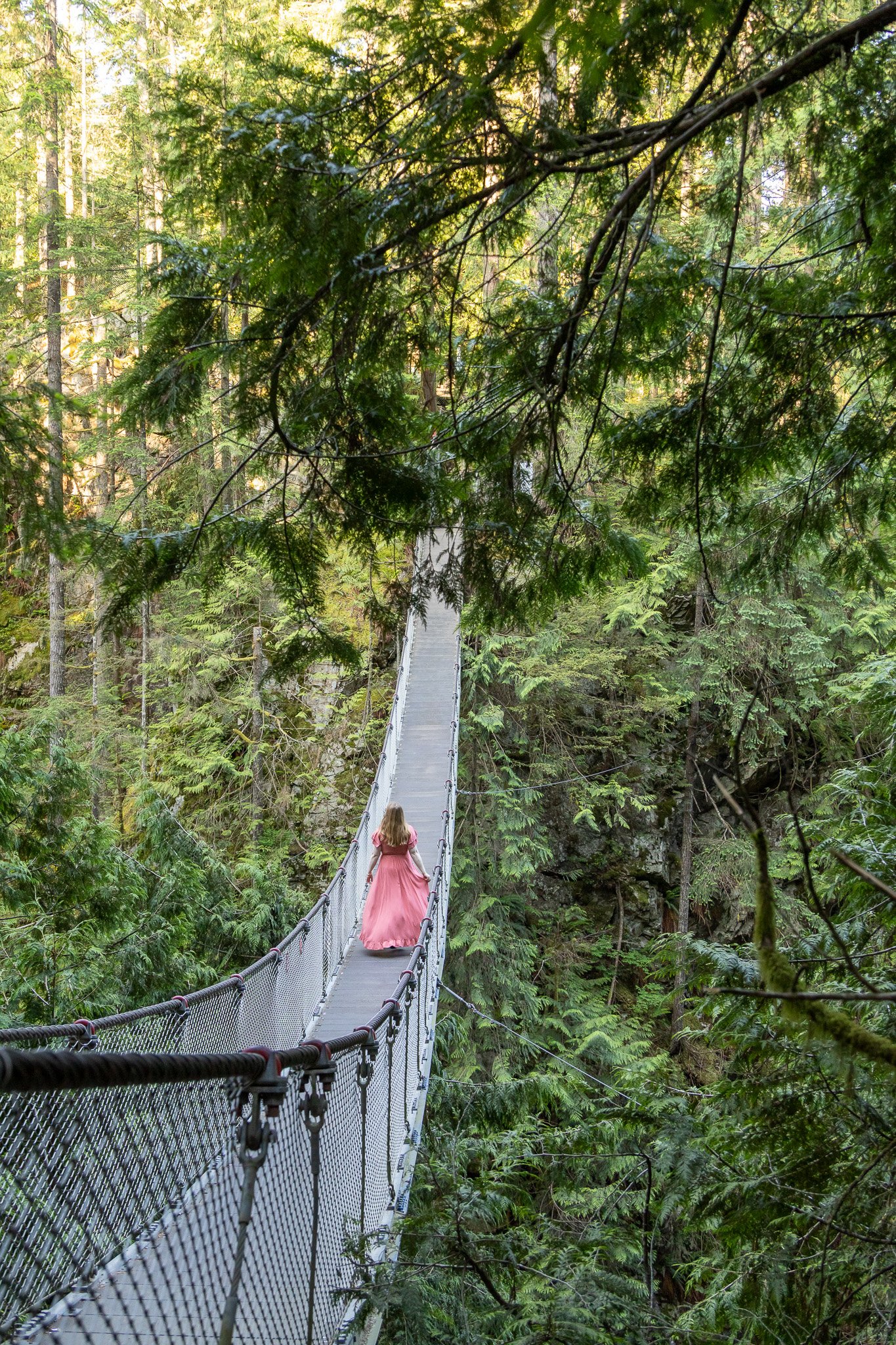 A woman in a long pink dress walking on a suspension bridge over a lush green forest.