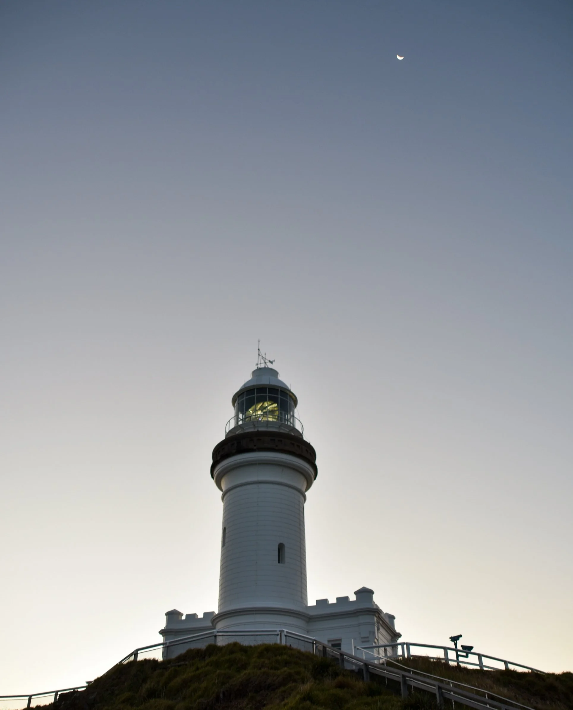 A lighthouse situated on a hill with a railing around it, against a evening sky that transitions from light to dark. The lighthouse has a white tower with multiple windows and a balcony near the top, topped by a lantern room with a light. A crescent moon is visible in the sky.