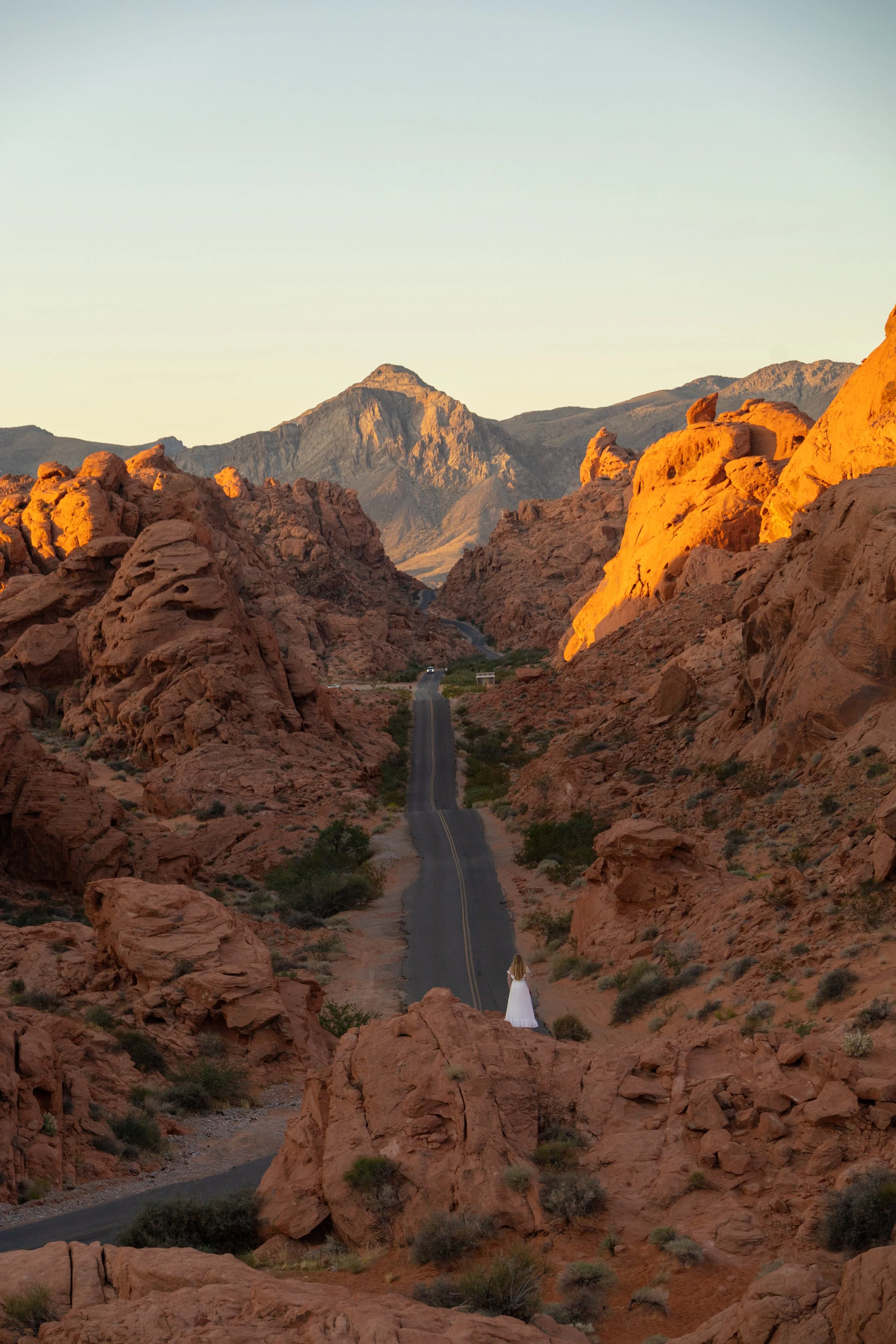 A woman in a white dress standing on rocky terrain in a desert landscape with large red rocks and mountains in the background, bathed in warm sunlight.
