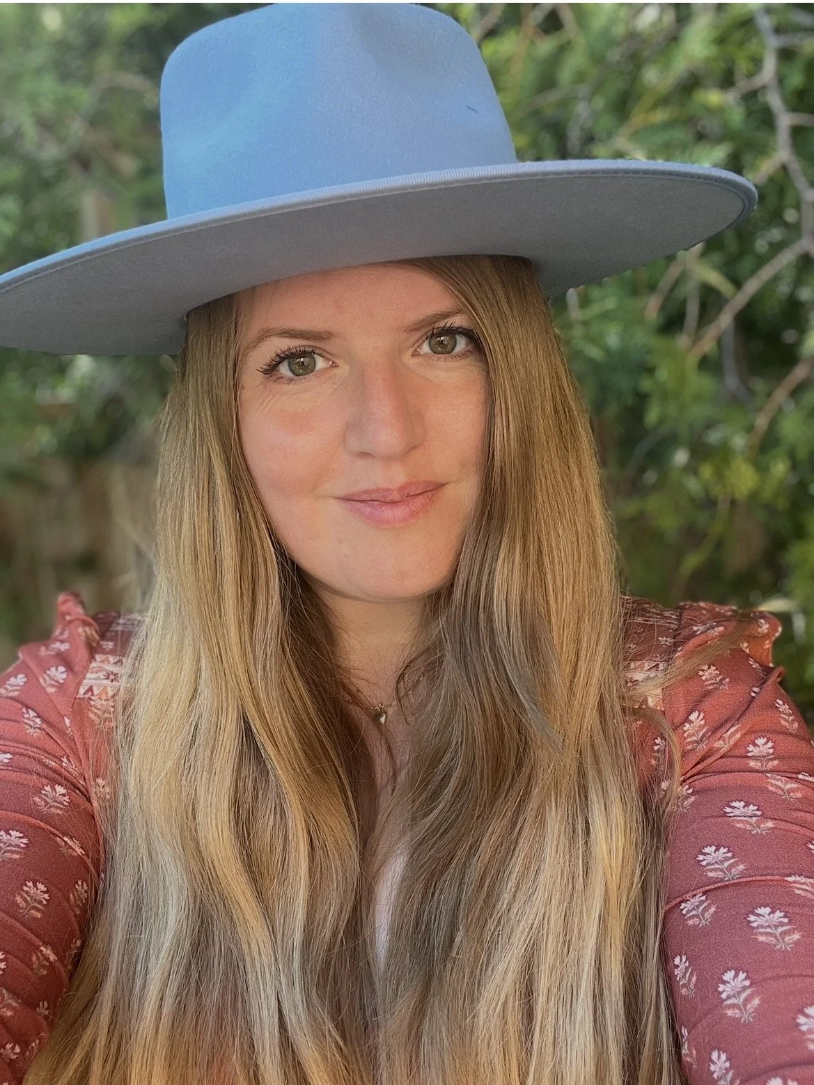 A woman with long blonde hair wearing a large light blue hat and a pink floral dress, standing outdoors with green foliage in the background.