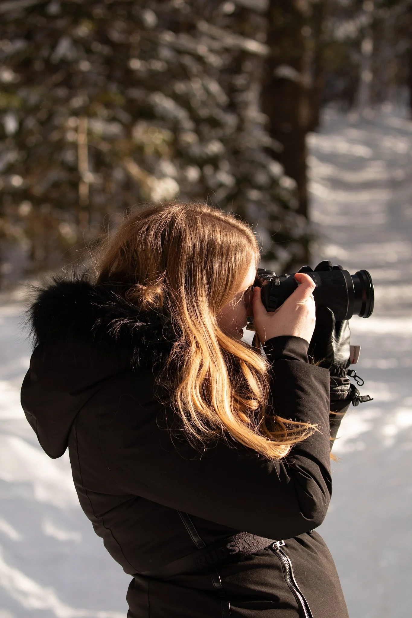 A woman with long, wavy blonde hair takes a photograph in a snowy landscape, wearing a black winter coat with a fur-lined hood.