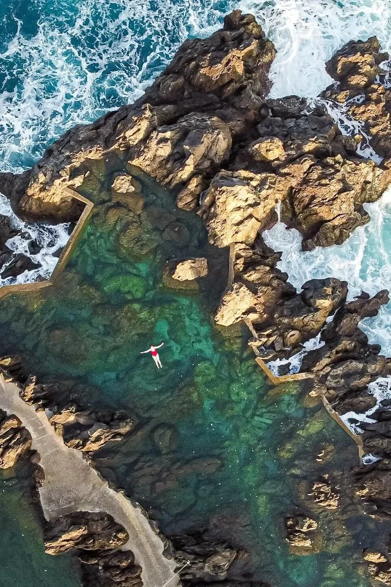 An aerial view of a rocky coastline with a natural ocean pool filled with clear water. A person is floating on their back in the pool, surrounded by rocks and waves crashing nearby.