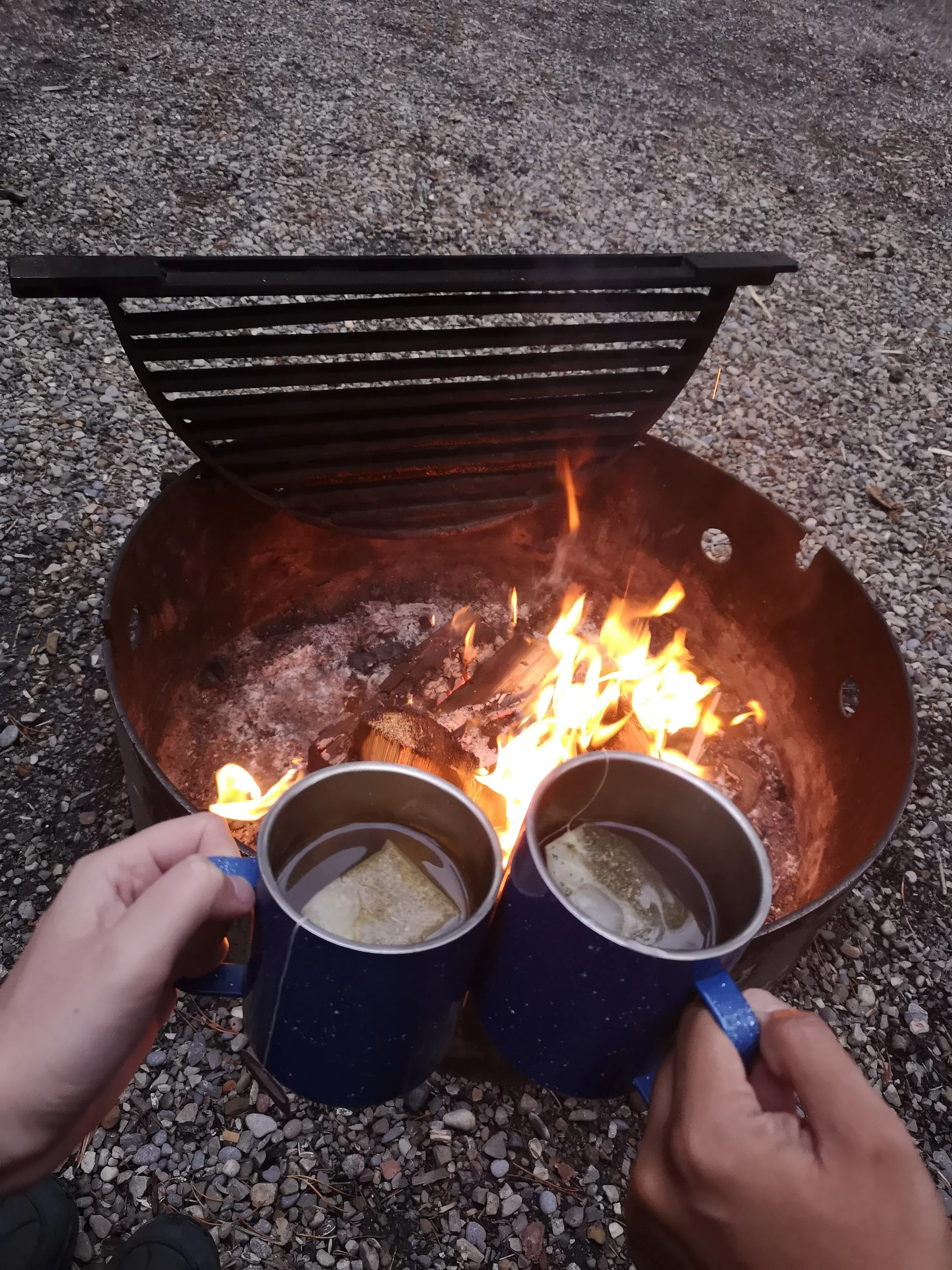 Two people holding mugs of hot drinks with marshmallows, sitting by a campfire with a grill, on rocky ground.