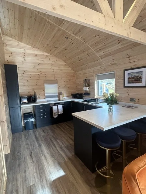 Kitchen inside a wooden cabin with curved ceiling, black cabinets, white countertops, a small window, a framed picture on the wall, and a kitchen island with seats.