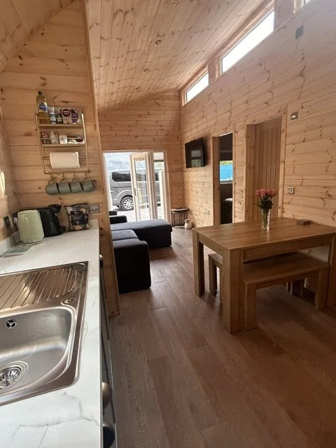 Interior of a cozy wooden cabin with a kitchen area, living room with couch, a wooden dining table with a flower vase, and a door leading outside.