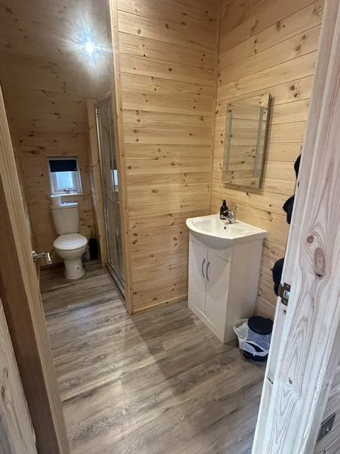 A small bathroom with light wood-paneled walls and laminate flooring. It features a white vanity with a mirror above, a small window with a black shade, and a toilet in a separate niche.