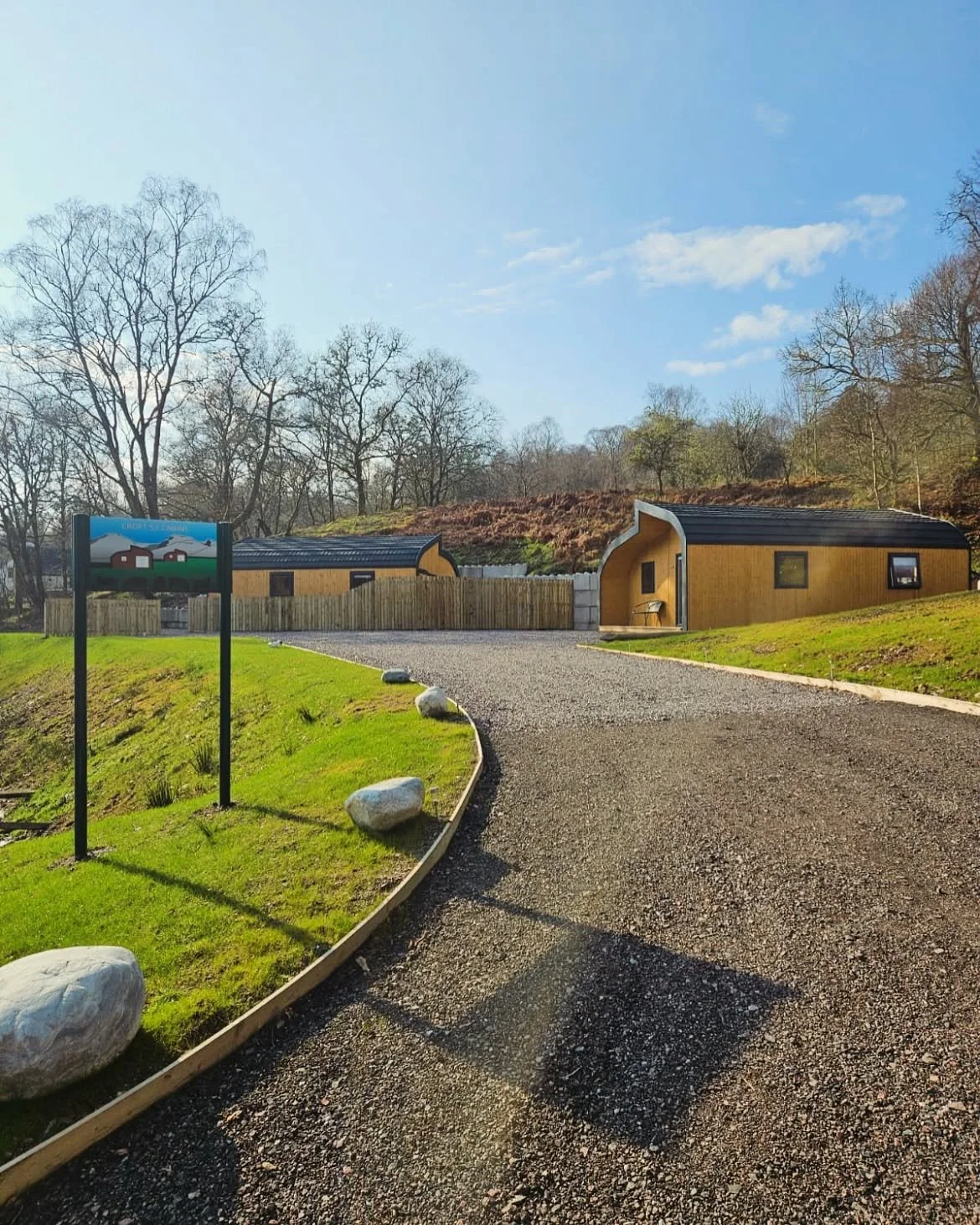 Modern building with curved roof, gravel driveway, green lawn, trees, and clear sky.