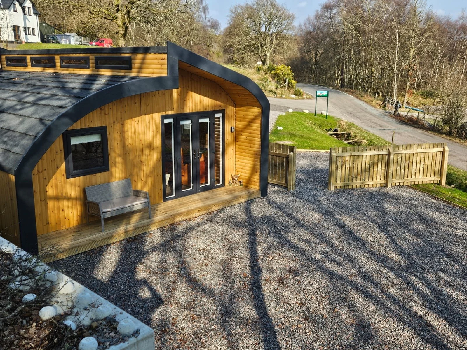 Wooden tiny house with curved roof, black trim, front door with glass panels, small porch with gray bench, wooden fence, gravel driveway, grassy area, trees and road in background.