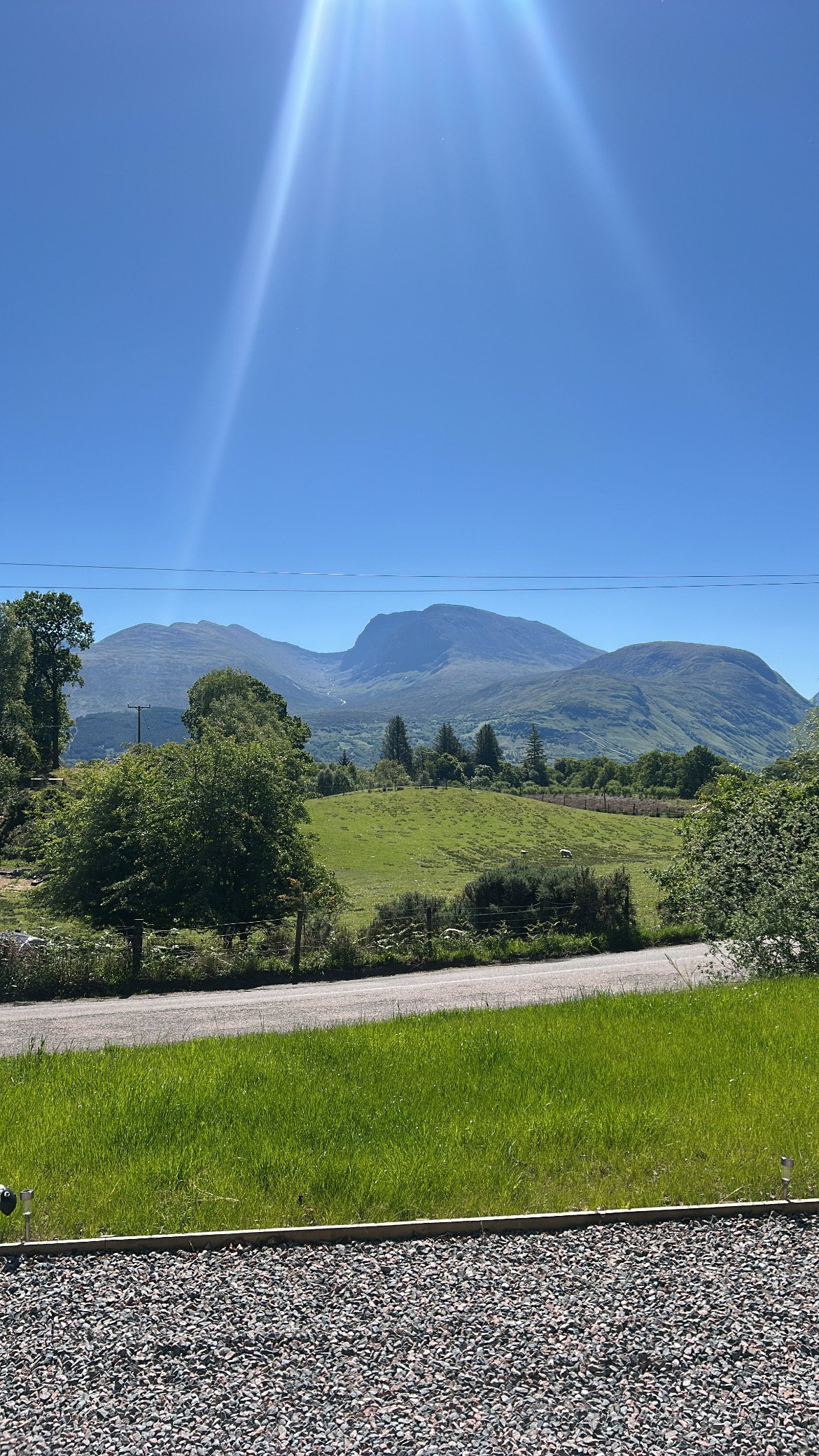 A scenic landscape with green grass, trees, and mountains under a clear blue sky with sunlight rays.