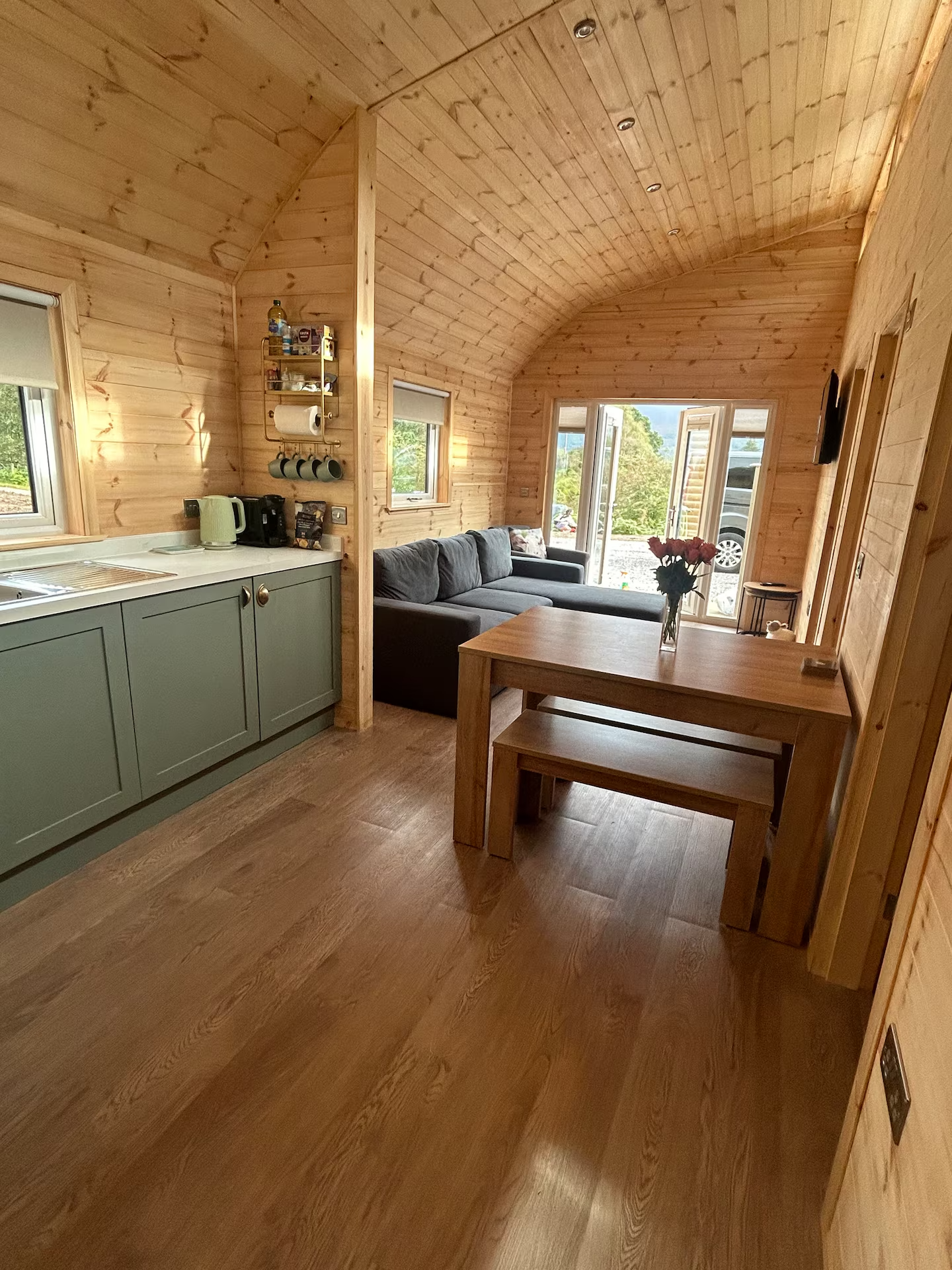 Interior of a cozy wooden cabin living area with a green kitchen cabinet, a gray sofa, a wooden dining table with a vase of flowers, and large windows showing outdoor scenery.