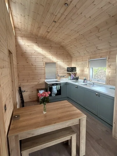 A cozy kitchen with light wood walls and ceiling, featuring a small dining table with a vase of pink flowers, gray cabinetry, a stove, microwave, and window letting in natural light.