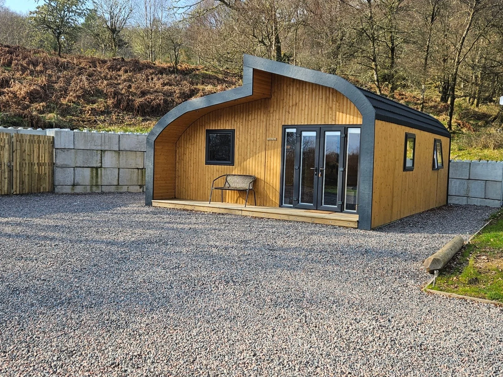 A modern tiny house with a curved roof, wooden walls, and large glass doors, situated on a gravel lot with a backdrop of trees and a hillside.