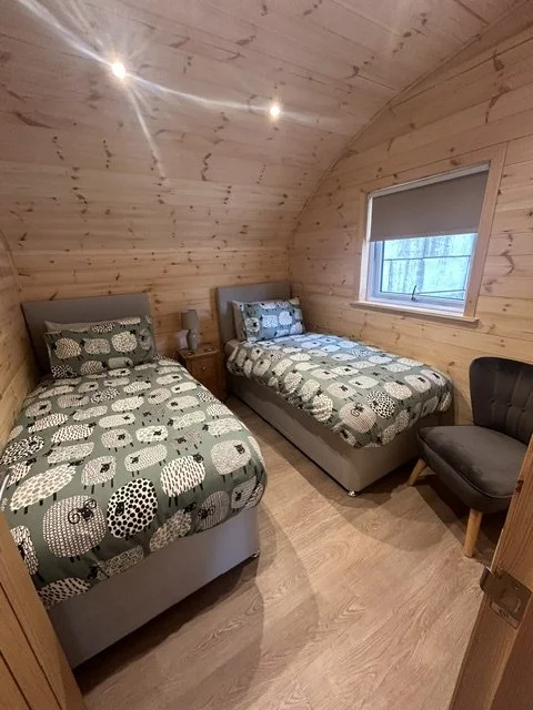 A cozy bedroom with wood-paneled walls and ceiling, two twin beds with animal-patterned bedding, a small nightstand with a lamp between the beds, a window with a blind, and a black armchair.