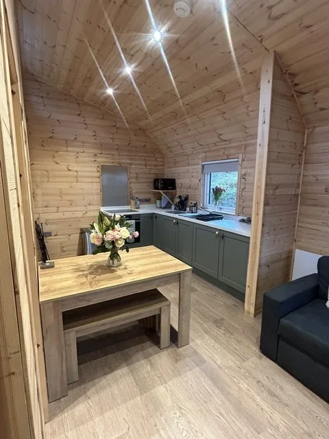 A cozy kitchen and dining area with wood-paneled walls and ceiling, green cabinets, a wooden dining table with a floral centerpiece, a black sofa, and a window with a flowerpot on the sill.