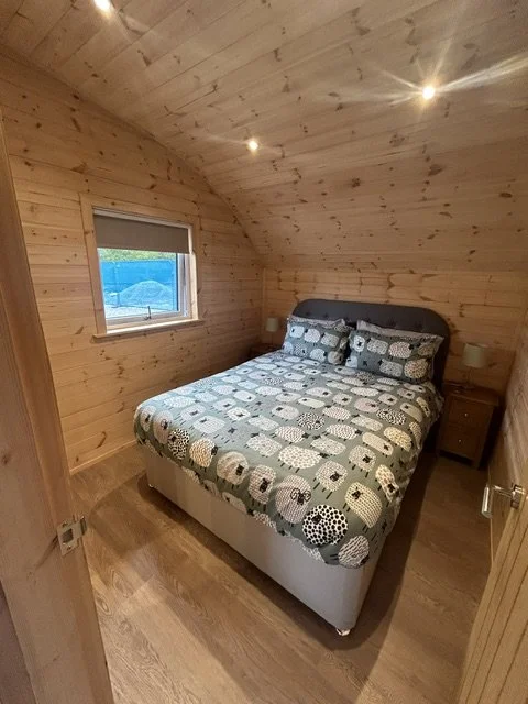 A cozy bedroom with wooden walls and ceiling, featuring a bed with a patterned quilt and two pillows, two small nightstands with lamps, a window with a view outside, and a wooden floor.