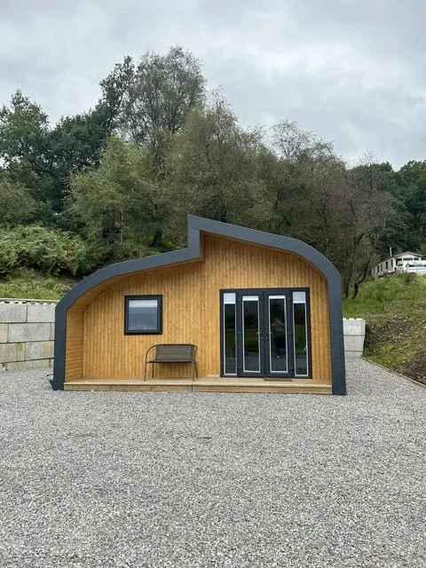 Small, modern wooden house with curved roof, set on a gravel lot, surrounded by trees.
