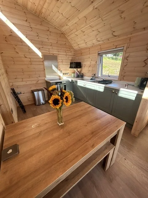 Small kitchen with wooden walls, a wooden dining table with a vase of sunflowers, gray cabinets, a window, and small appliances on the counter.