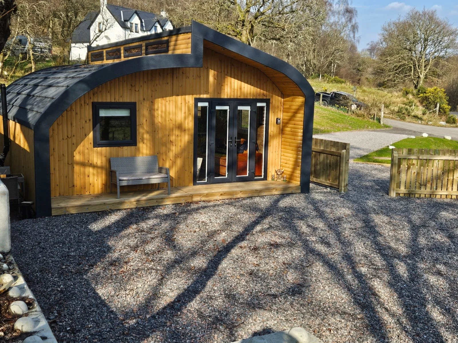 A small wooden cabin with a curved roof, black framing around the door and window, and a small porch with a bench, situated in a rural area with gravel ground and trees in the background.