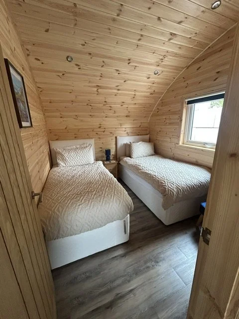 A cozy bedroom with two beds, quilted beige bedding, wooden walls and ceiling, a window, and a small nightstand between the beds with a lamp.