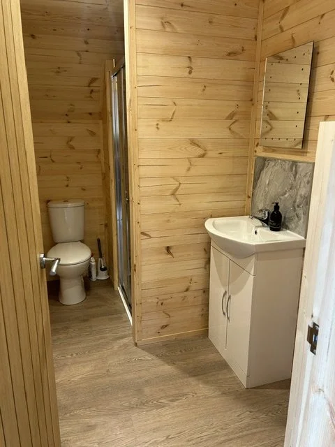 Small bathroom with wooden walls, a white toilet in the background, a small white sink with a mirror above it, and a wood and marble wall behind the sink.