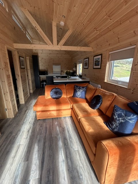 Interior of a cozy wooden cabin with a bright orange sectional sofa, dark blue pillows, wood-paneled ceiling and walls, a window showing greenery outside, and a kitchen area in the background.