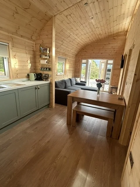 Interior of a cozy wooden cabin with a kitchenette, a dining table, and a seating area with a sofa near large windows showing greenery outside.
