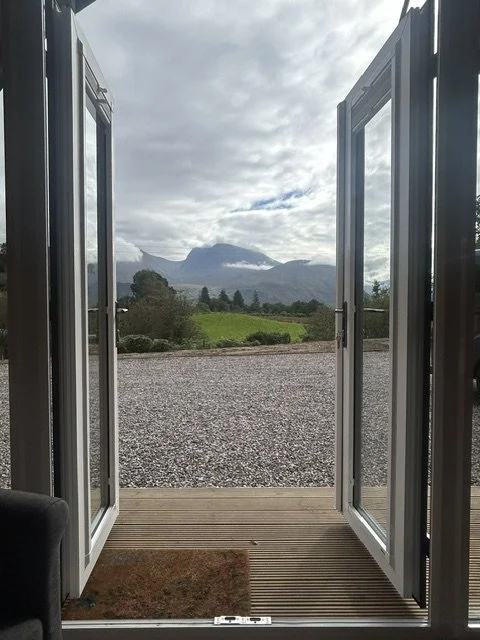 Open glass doors showing a gravel pathway, green fields, and mountains under a cloudy sky.