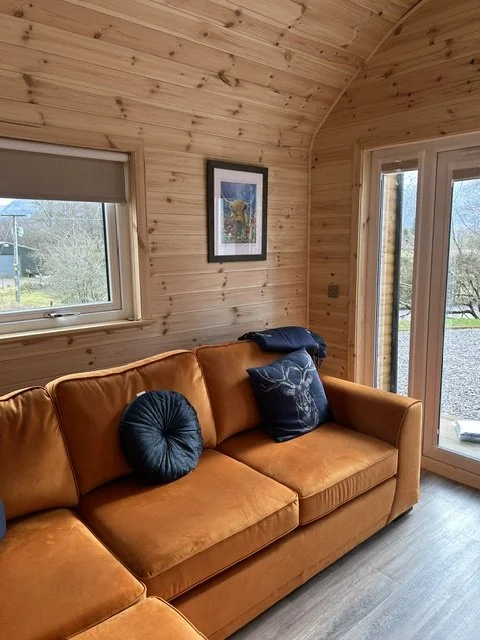 Interior of a cozy wooden room with a large orange sofa, two decorative pillows, and framed artwork on the wooden wall. There are windows and a glass door showing an outdoor view of trees and grass.