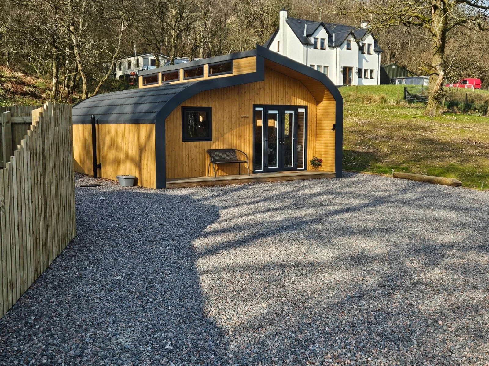 A small, modern wooden cabin with a curved roof and black trim, situated on a gravel yard with a wooden fence on the left. Behind the cabin are trees and a white house on a hill.
