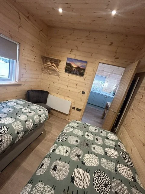 Two beds with patterned bedspreads in a wooden room, window with blinds, small black couch, doorway leading to a kitchen area.