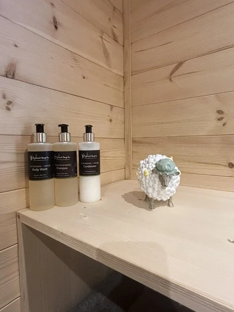 Three bottles of body wash and shampoo alongside a decorative ceramic sheep figurine on a wooden shelf in a corner with light wood paneling.