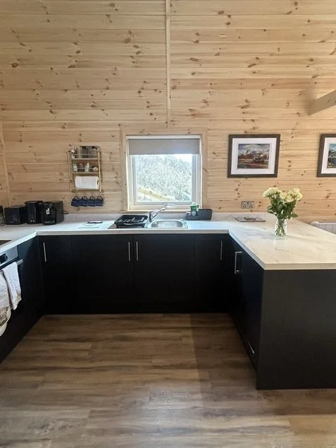 Kitchen with black cabinets, marble countertops, wood-paneled walls, a window above the sink, framed pictures on the wall, a vase of flowers, and various small appliances on the counter.