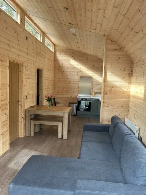 Wood-paneled living room with a blue sectional sofa, a wooden dining table with matching benches, a vase with pink flowers, and a compact kitchen area with a stove and microwave, illuminated by natural light through high horizontal windows.