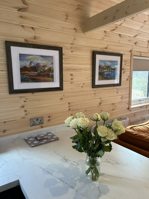 A vase with white roses placed on a white countertop in a room with wooden walls and two framed paintings hanging on the wall.