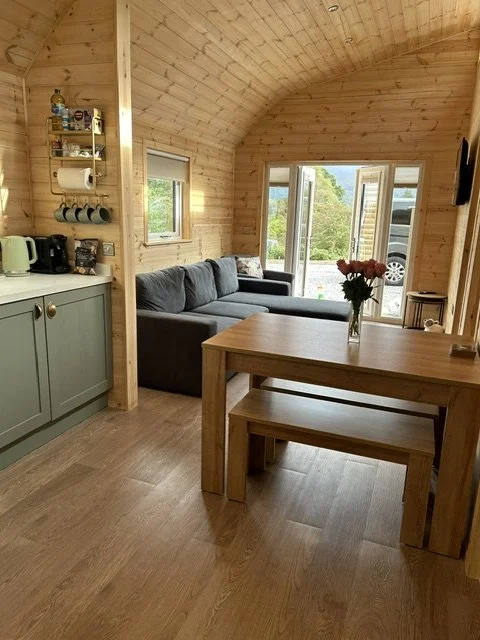 Interior of a cozy wooden cabin with a dining table, a sofa, kitchen area, and large windows showing greenery outside.