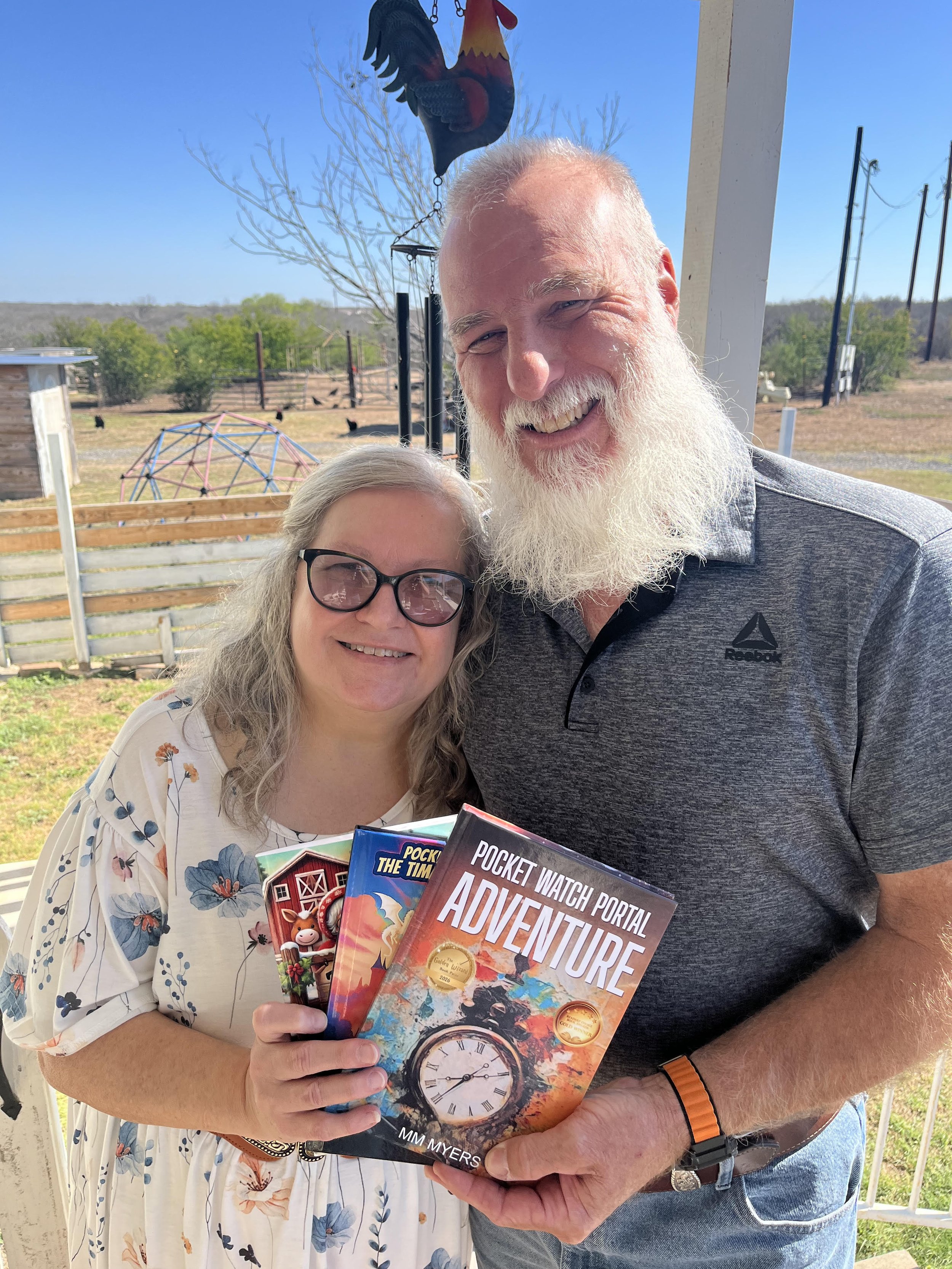 A woman with glasses holding a book titled 'Pocket Watch Portal Adventure' standing next to a man with a white beard and glasses, holding several colorful children's books at an outdoor location with a clear blue sky and a rooster-shaped weather vane in the background.